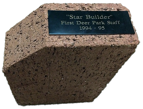 Photograph of the brick doorstop described by Donna Ford. The color is a light reddish-brown. A small plaque, affixed to the top, reads Star Builder, First Deer Park Staff, 1994 to 1994. The brick is pentagonal, not rectangular, in shape. 