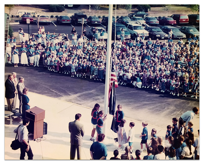 Color photograph taken from the roof of Deer Park Elementary School during its dedication ceremony. We are looking down at a crowd of students gathered in front of the school as another group of students is participating in a flag raising ceremony.